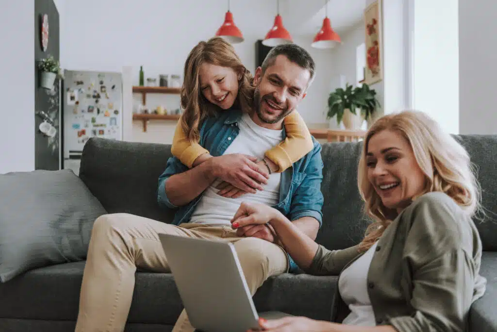 Cute little girl hugging her father from behind while he sitting on couch and holding hand of his wife. They looking at notebook display and smiling